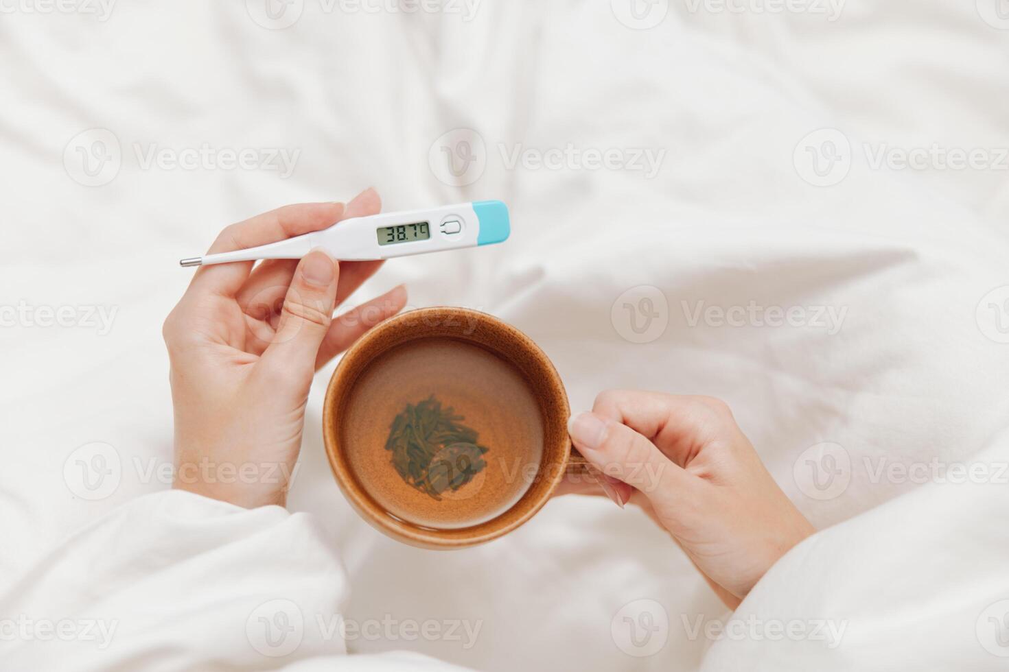 Sick person lying in bed holding a thermometer and a cup of tea, with another thermometer on the photo