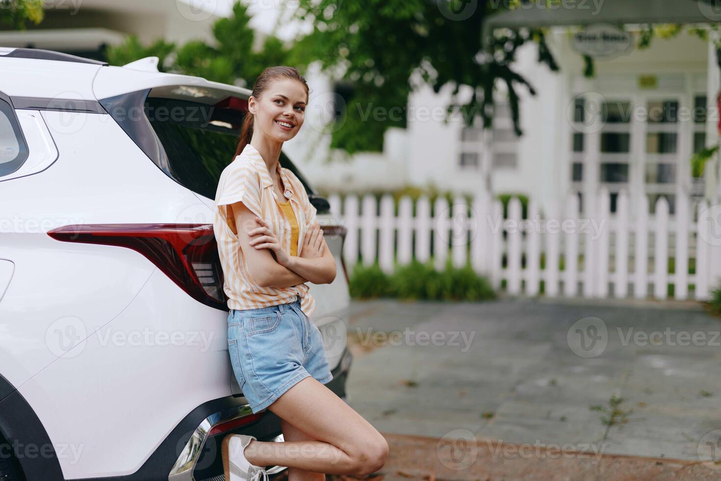 A happy young woman stands casually against a white SUV in a sunny outdoor setting She wears a photo