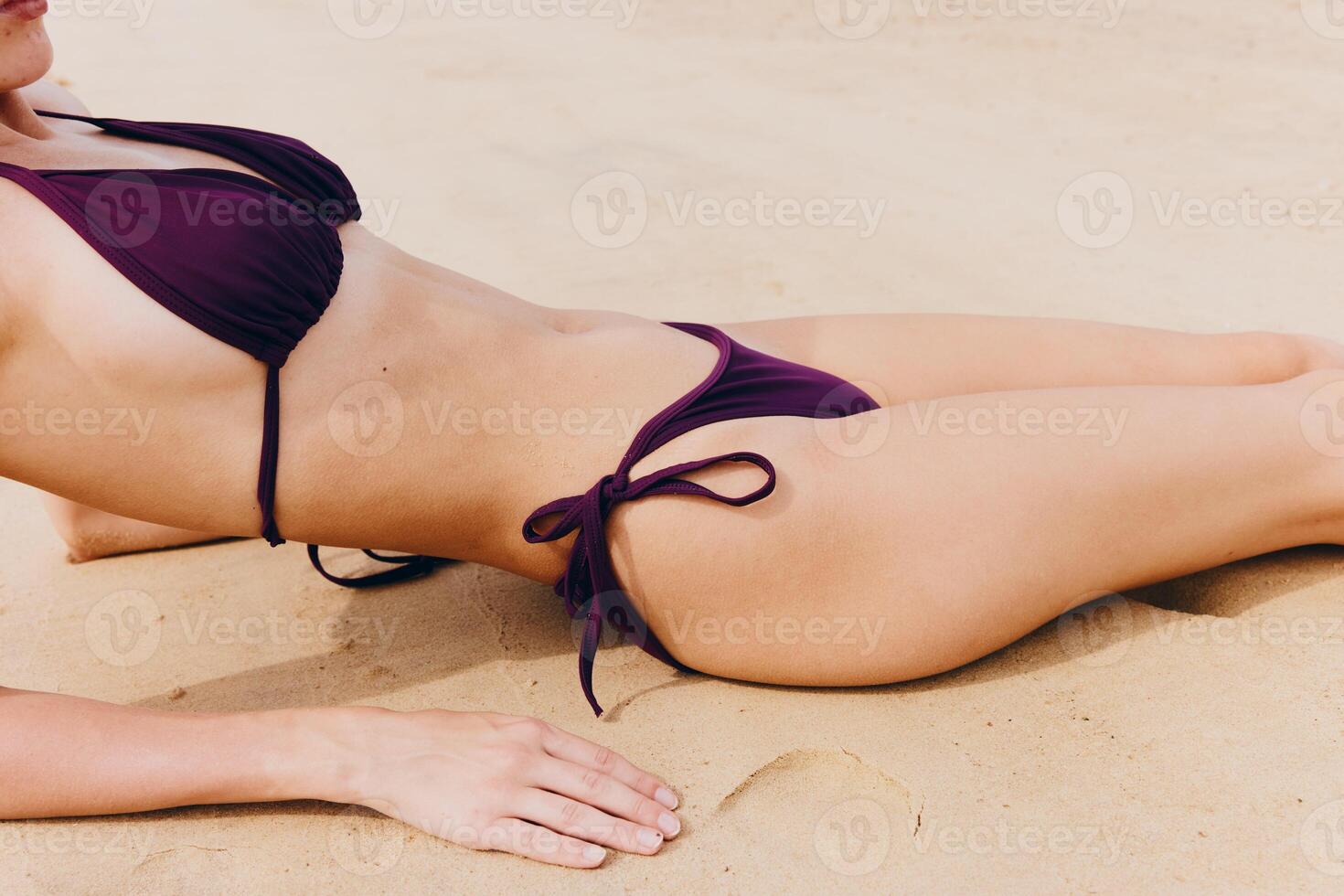Woman in purple bikini lying on sandy beach, hands on hips and head tilted, looking directly at the photo