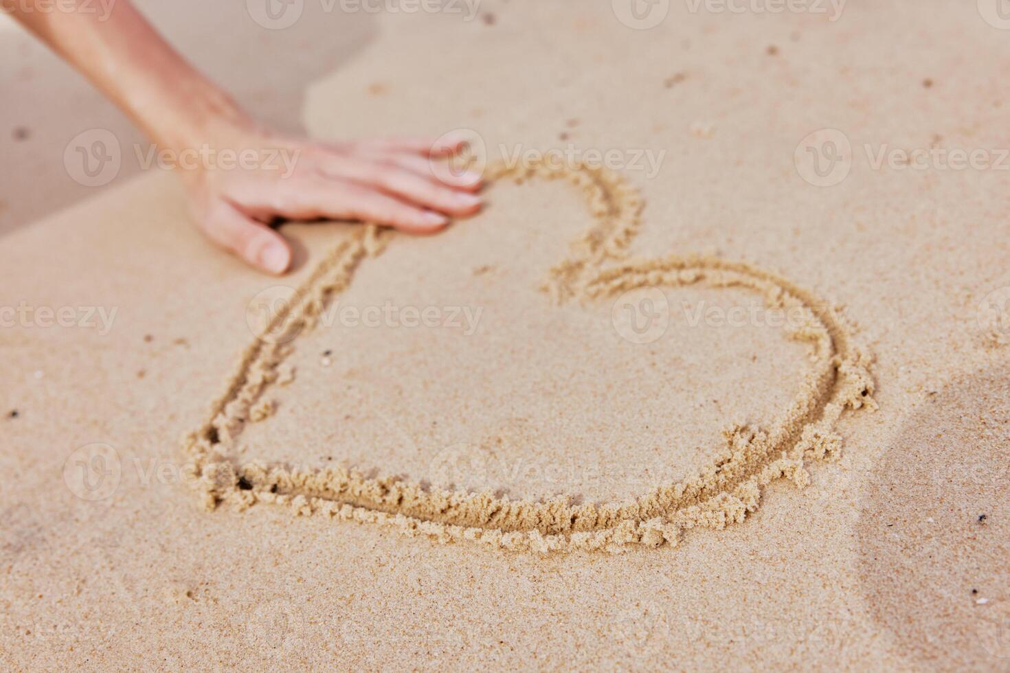 Person drawing heart in sand on beach, with another hand reaching out to touch it. romantic and photo