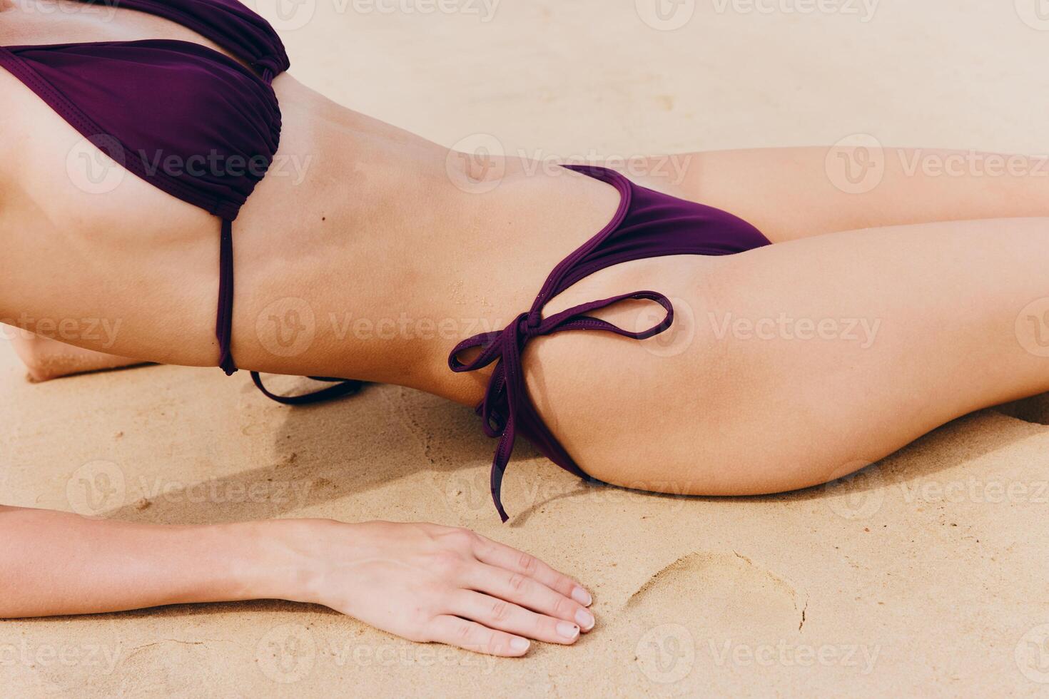 Woman in purple bikini lounging on sandy beach with legs apart, hands on hips, and head tilted to photo