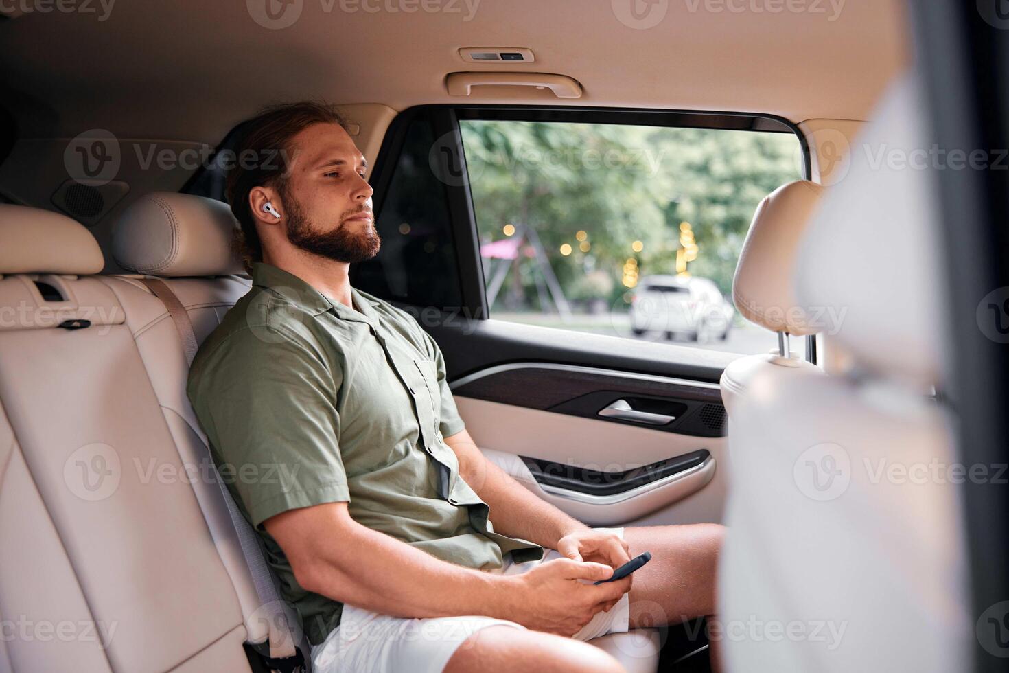 A man in a light green shirt and white shorts sits calmly in a car s backseat, wearing wireless photo