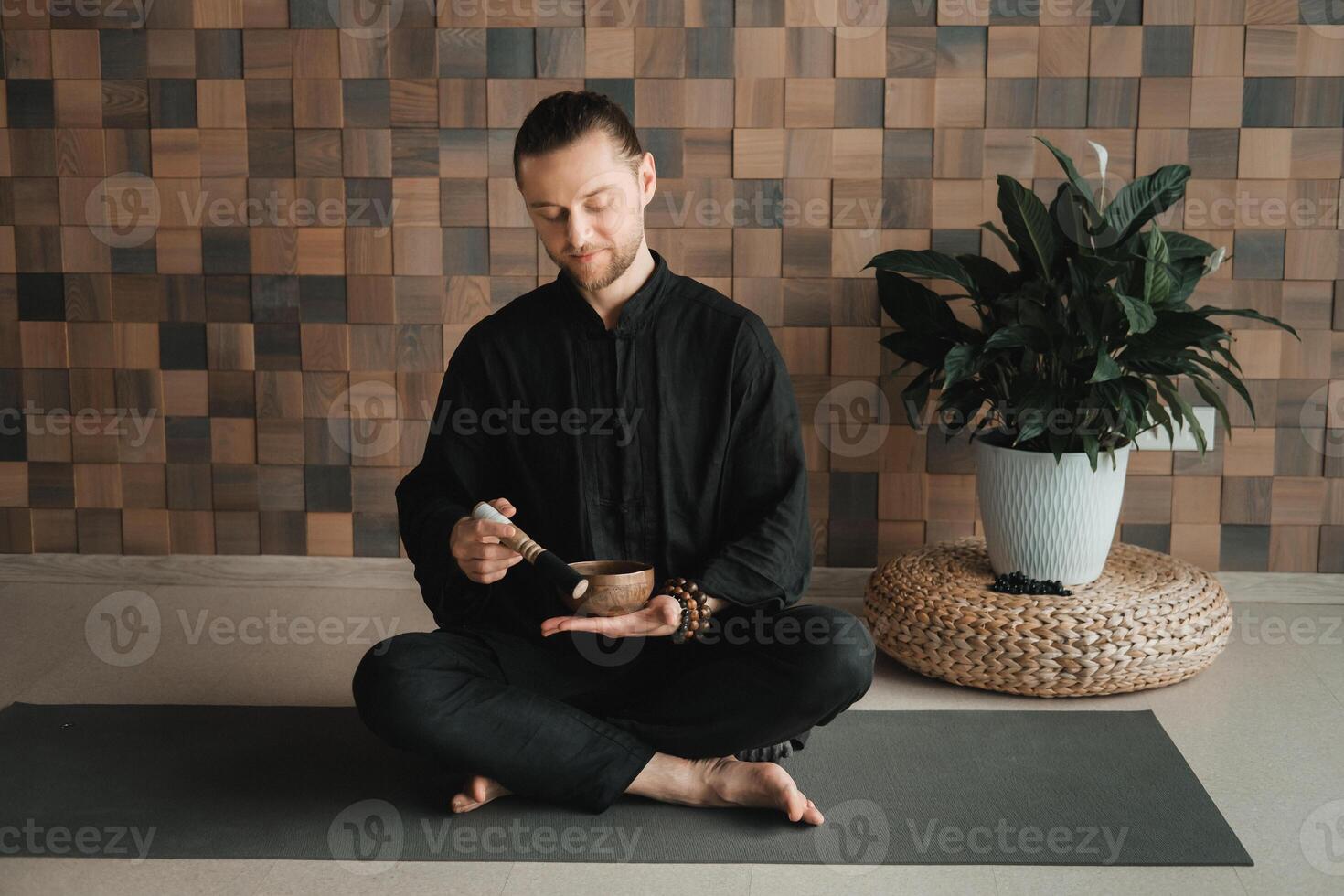 Portrait of a young man in a black kimono, holding a Tibetan bowl in his hands, sitting in a lotus position on a gym table in the interior photo