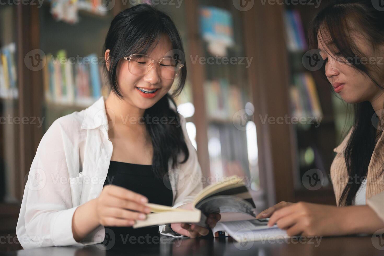 Two women are sitting at a table in a library, one of them reading a book photo
