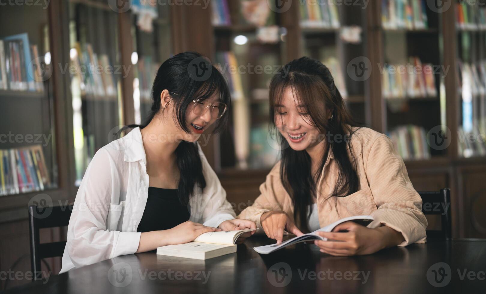 Two women are sitting at a table in a library, looking at a book photo