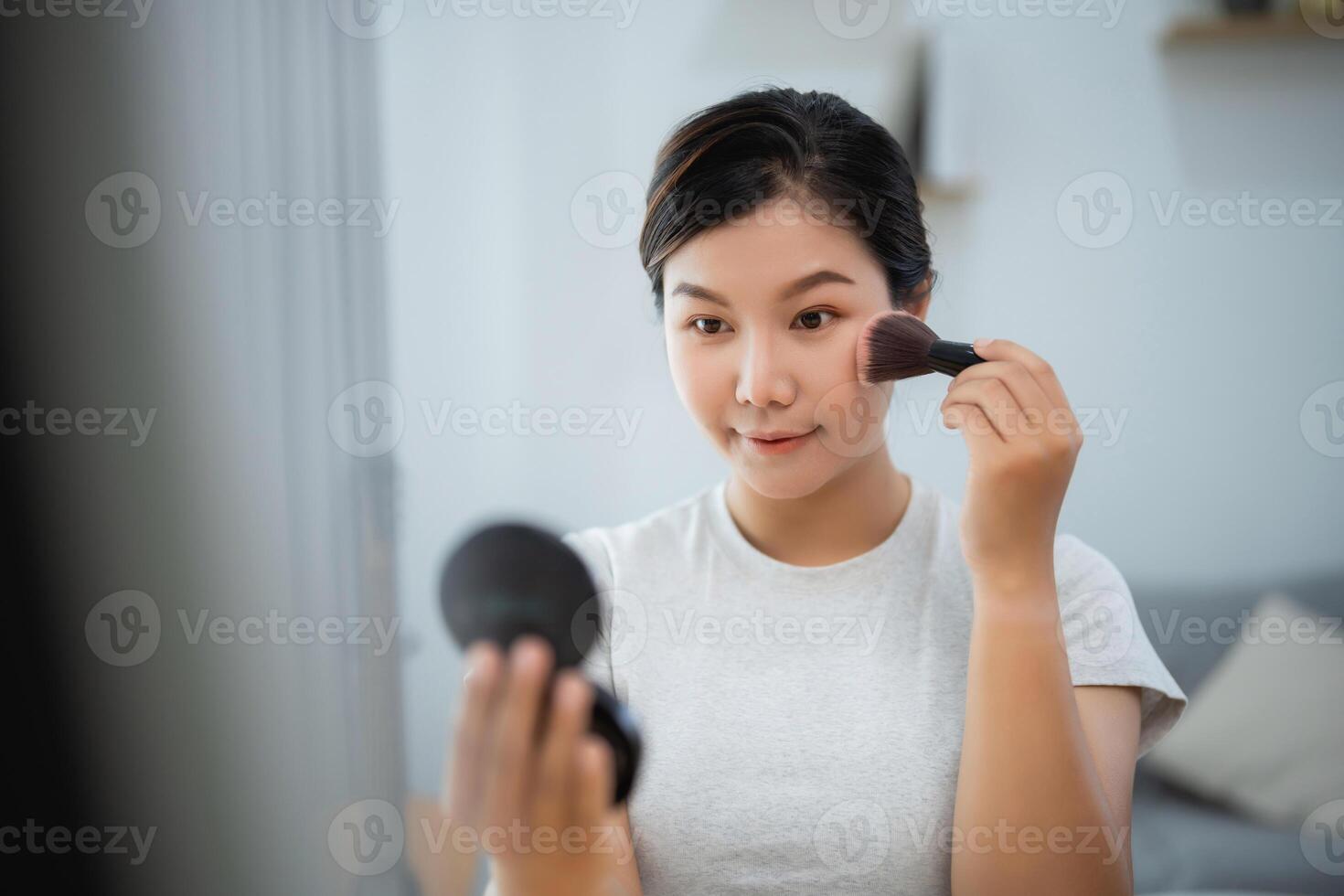 Asian woman is applying makeup in front of a mirror. She is holding a brush and is looking at herself in the mirror. Concept of self-care and personal grooming photo
