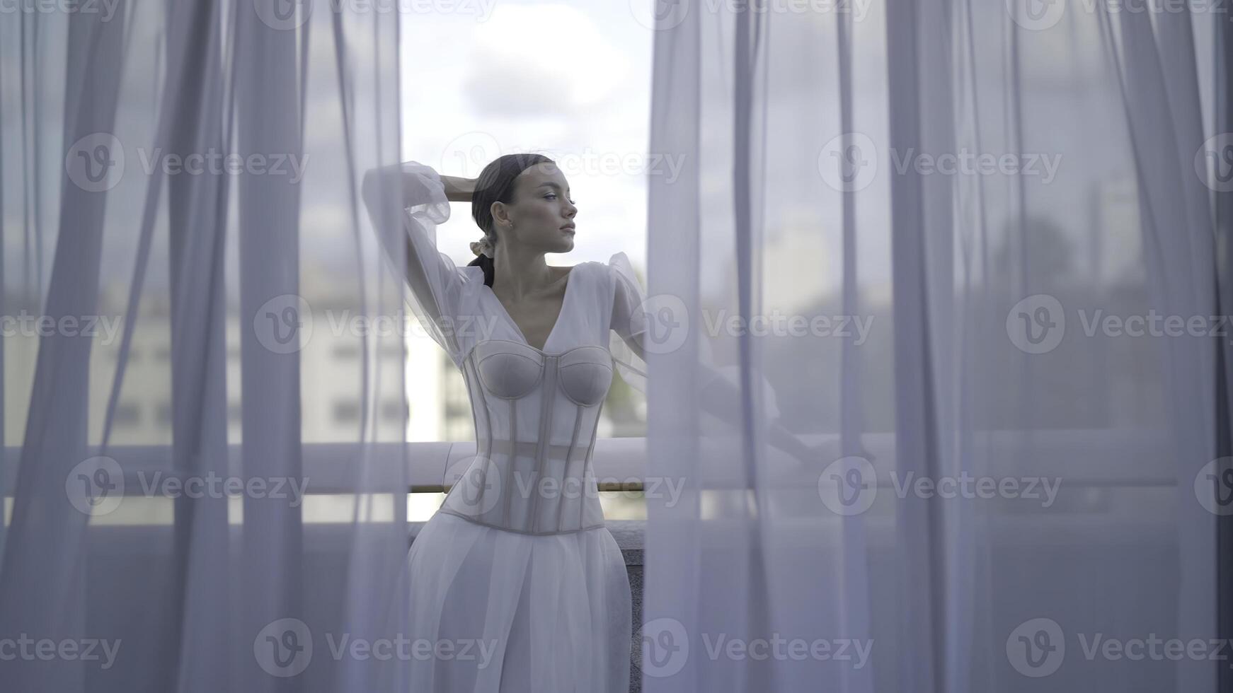 Beautiful woman posing in white dress with corset. Action. Elegant woman inwhite dress poses on terrace. Beautiful view through tulle of posing woman in white dress on terrace photo