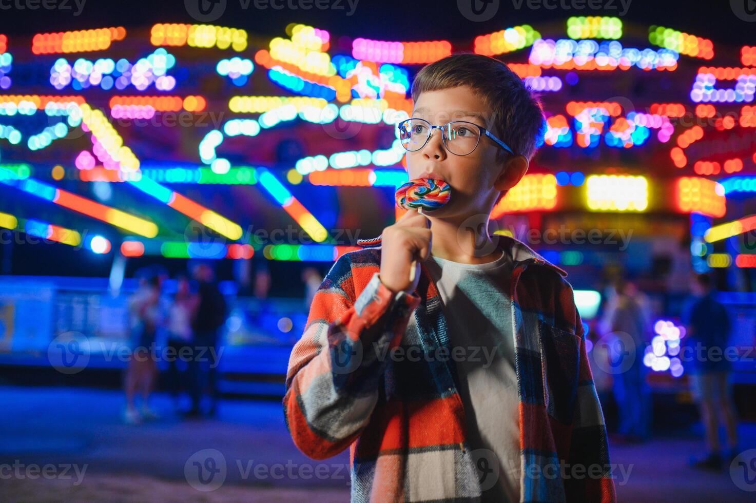 A cute caucasian boy eating a colorful lollipop standing against the background of a carousel in the evening at an amusement park or circus photo