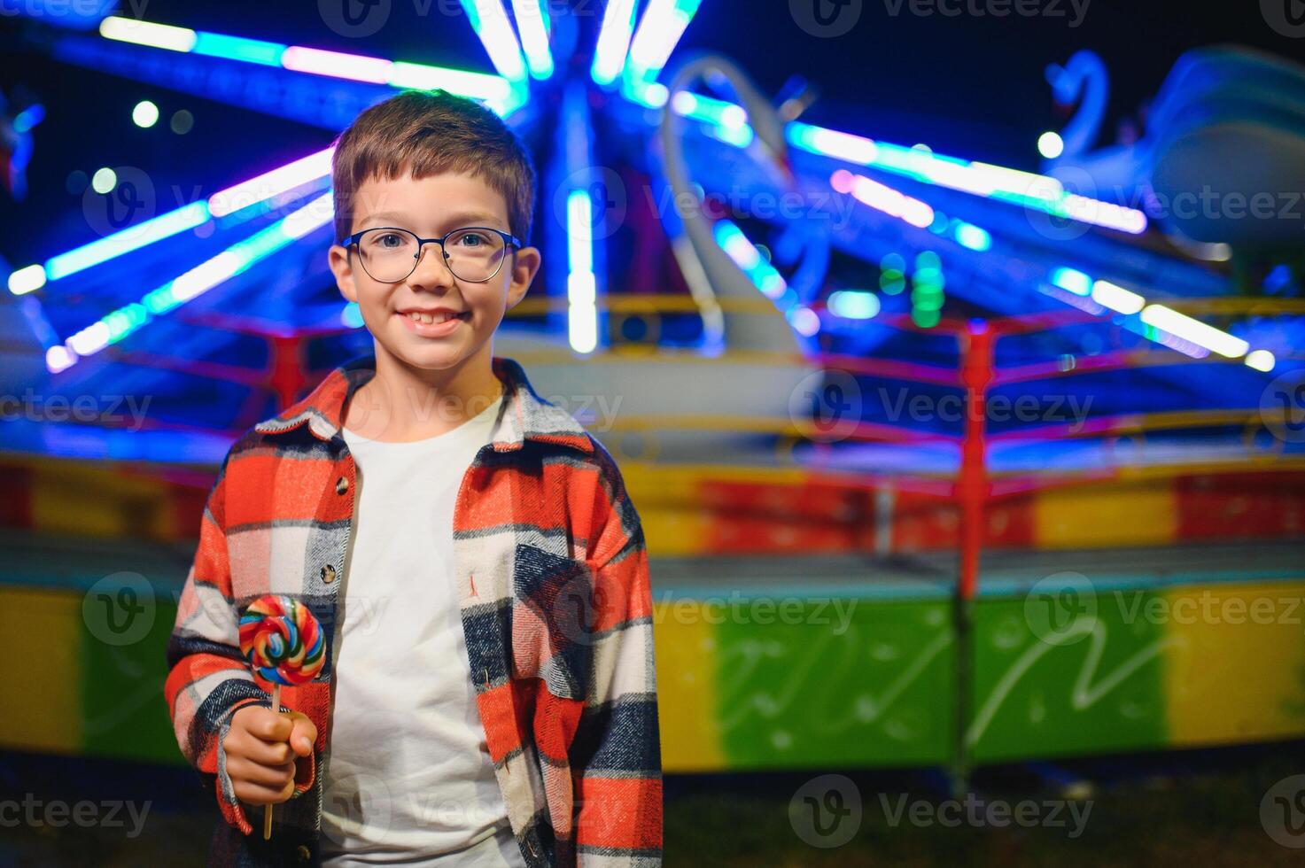 A cute caucasian boy eating a colorful lollipop standing against the background of a carousel in the evening at an amusement park or circus photo