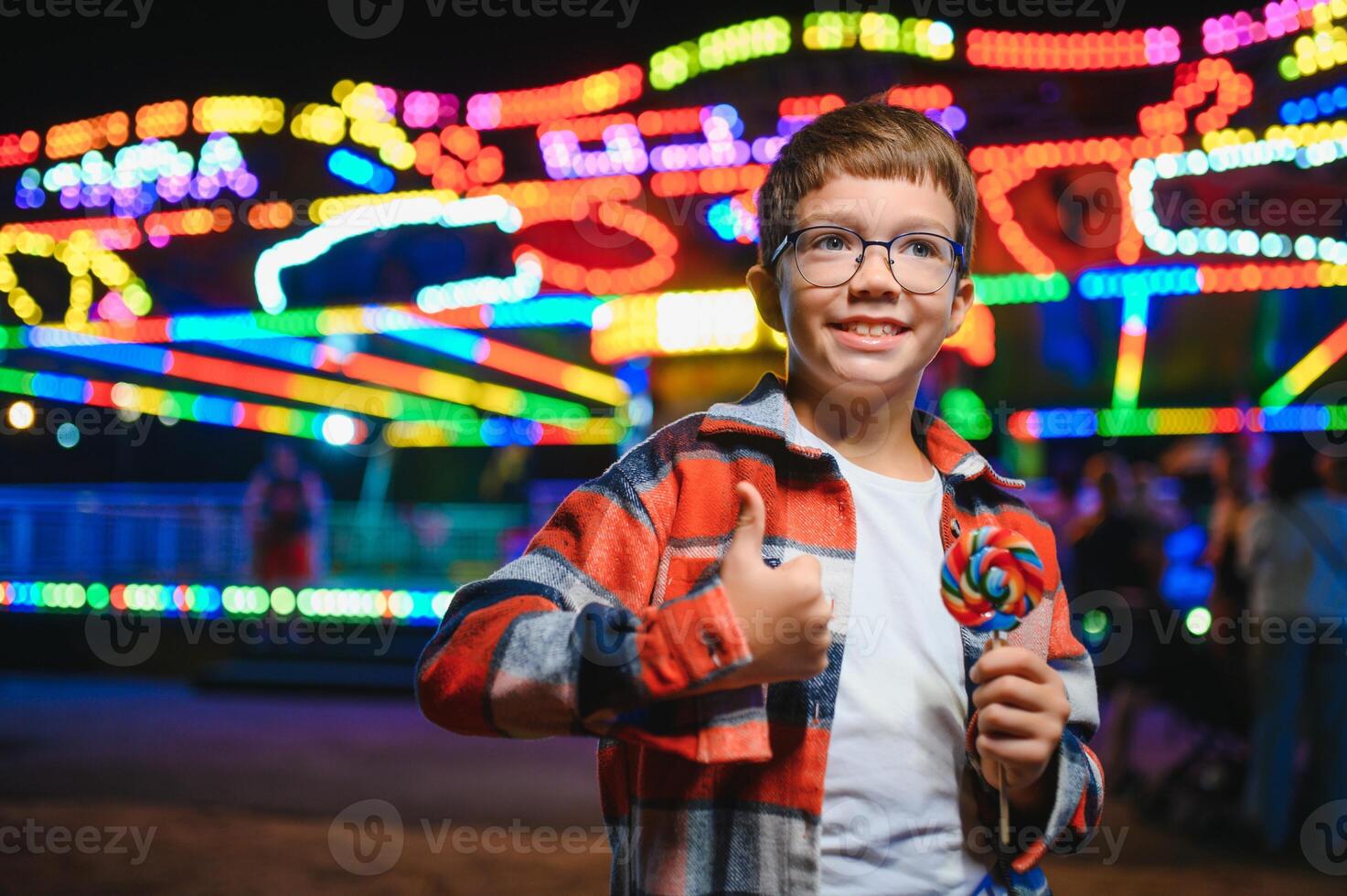 A cute caucasian boy eating a colorful lollipop standing against the background of a carousel in the evening at an amusement park or circus photo