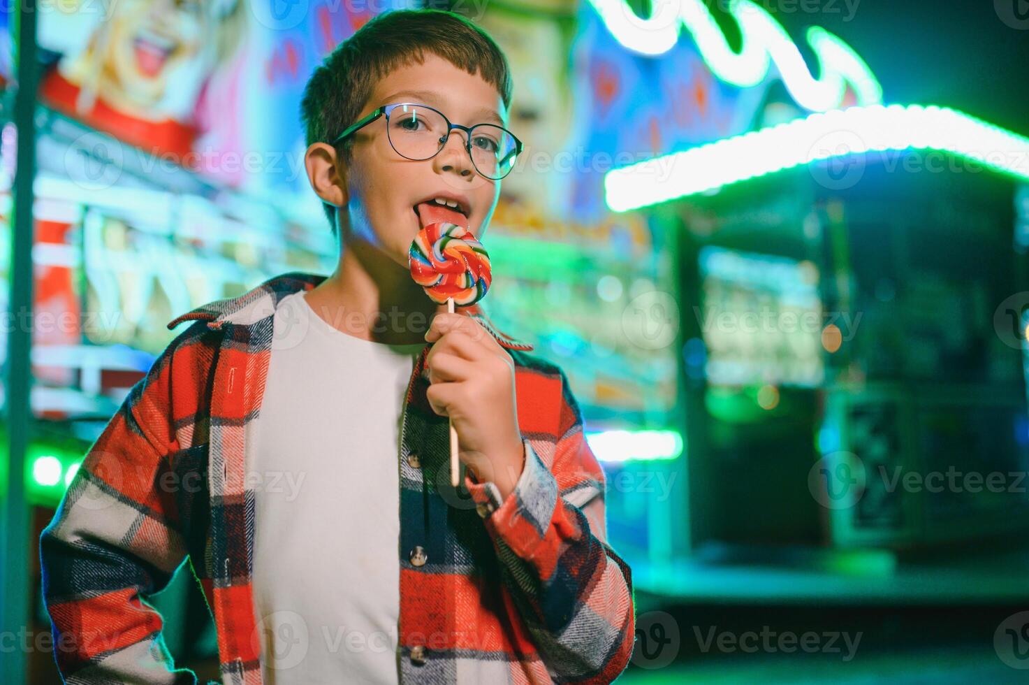 A cute caucasian boy eating a colorful lollipop standing against the background of a carousel in the evening at an amusement park or circus photo