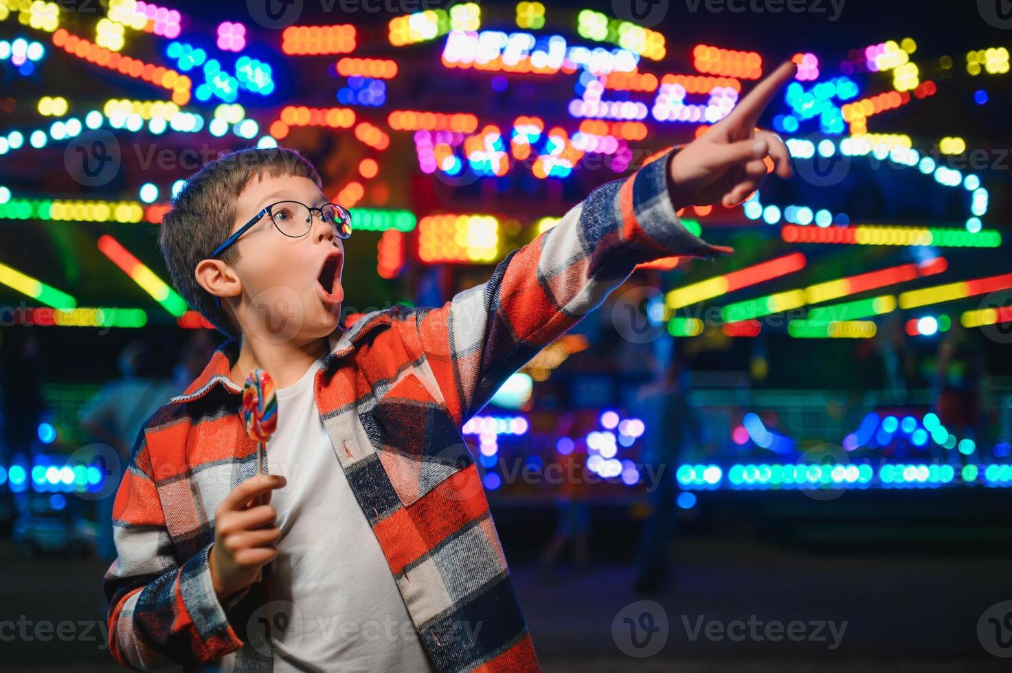 A cute caucasian boy eating a colorful lollipop standing against the background of a carousel in the evening at an amusement park or circus photo