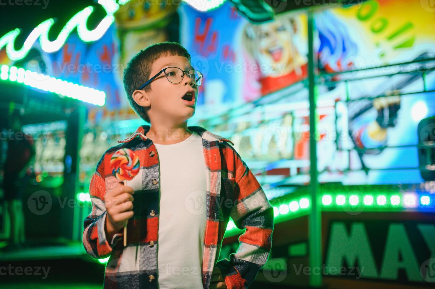 A cute caucasian boy eating a colorful lollipop standing against the background of a carousel in the evening at an amusement park or circus photo