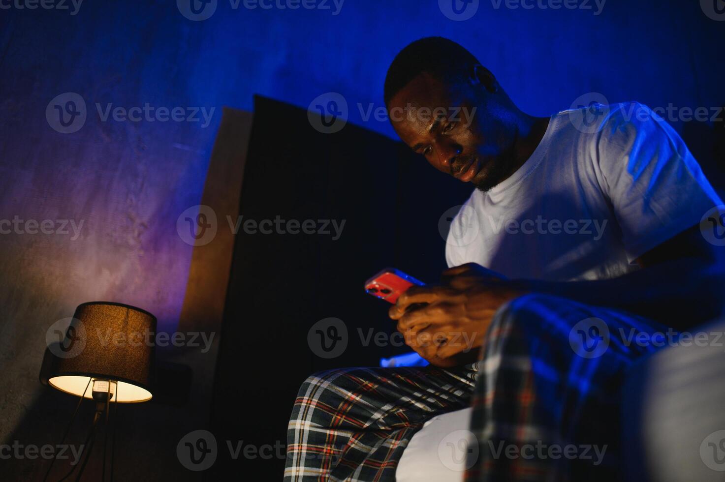 Low angle portrait of adult African-American man using smartphone while sitting on bed at night. Problems with insomnia. photo