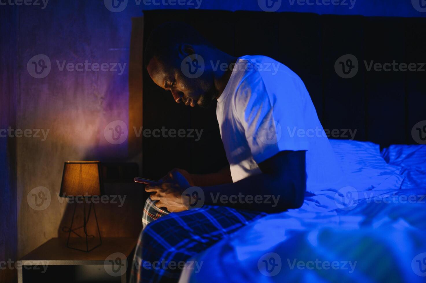 Low angle portrait of adult African-American man using smartphone while sitting on bed at night. Problems with insomnia. photo