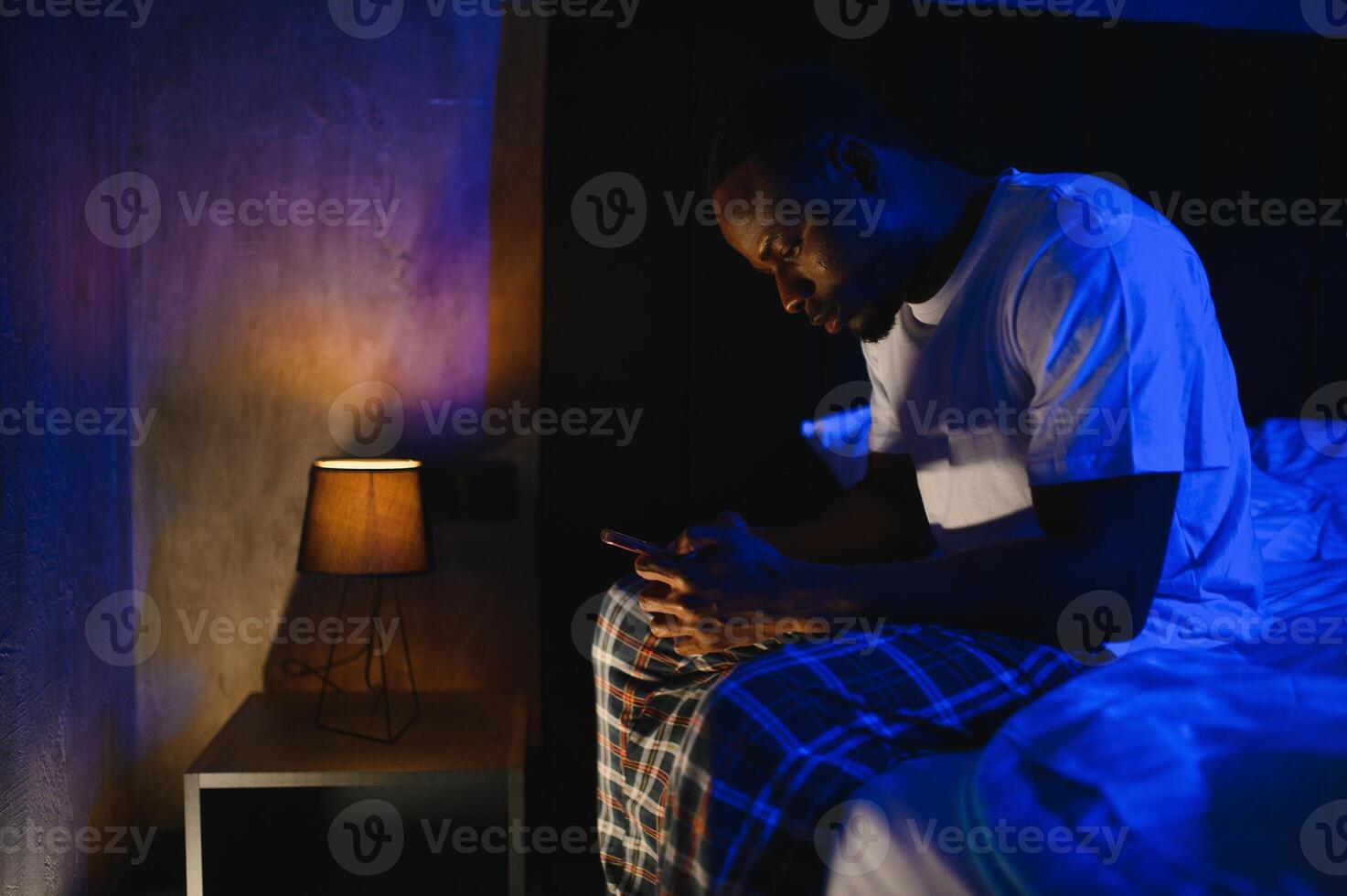 Low angle portrait of adult African-American man using smartphone while sitting on bed at night. Problems with insomnia. photo