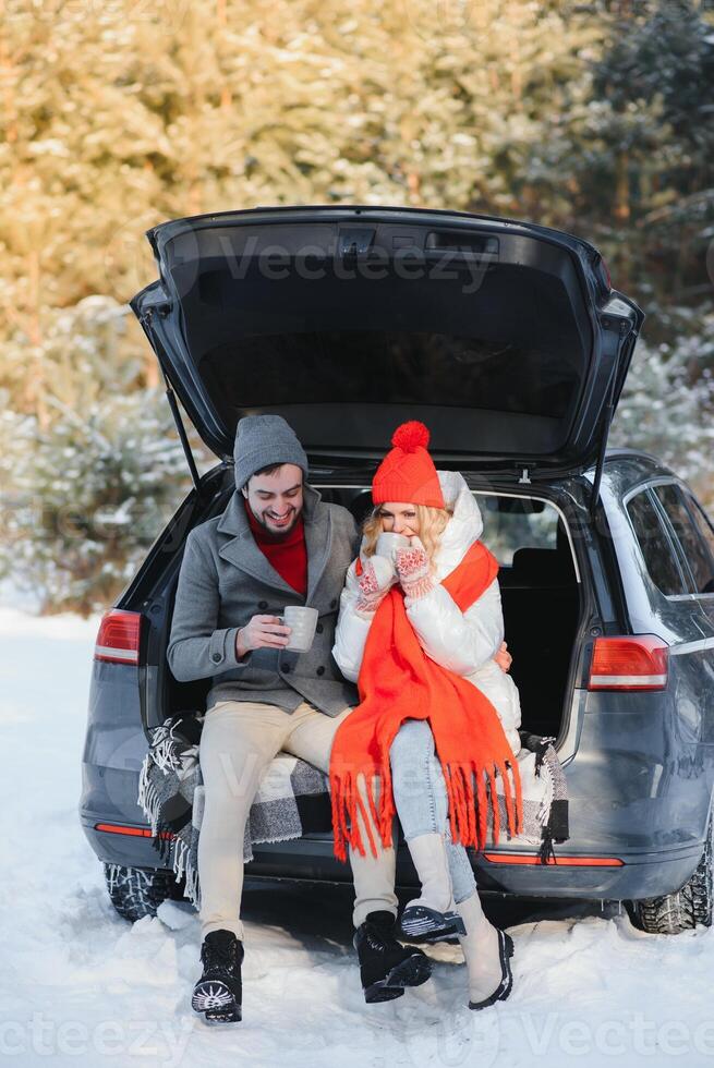 Happy Young couple is sitting in the back trunk of car in winter forest and drinking hot tea from thermos. Winter time photo