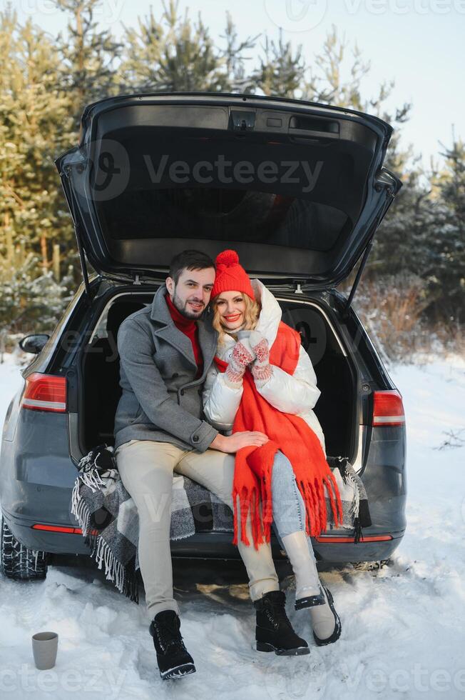 Couple in love sitting in car trunk drinking hot tea in snowy winter forest and chatting. People relaxing outdoors during road trip. Valentines day photo