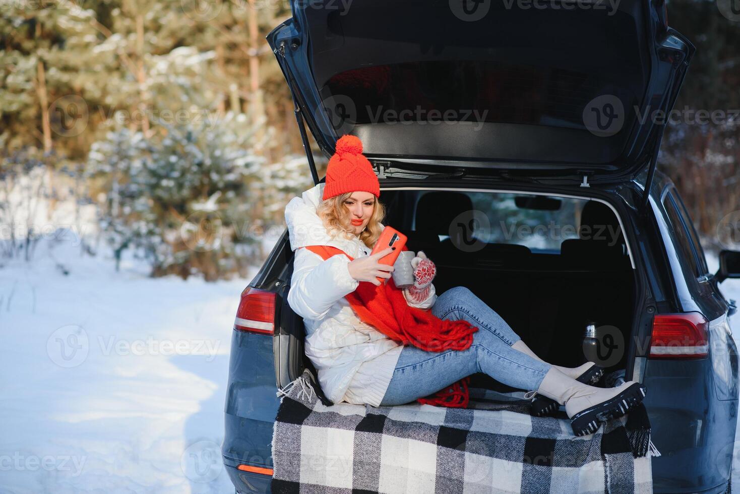 young woman sitting in car trunk drinking warm tea and takes a selfie at winter snowed day. photo