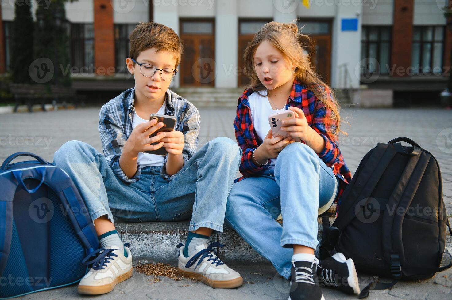 Two school little students small boy and girl sitting on stairs near modern school building. Pretty friends searching internet on smartphone and showing to friend. photo