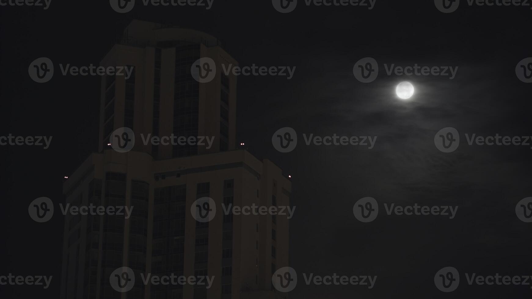 Night cityscape with a high building on dark sky background. Stock footage. Full moon on dark cloudy sky. photo