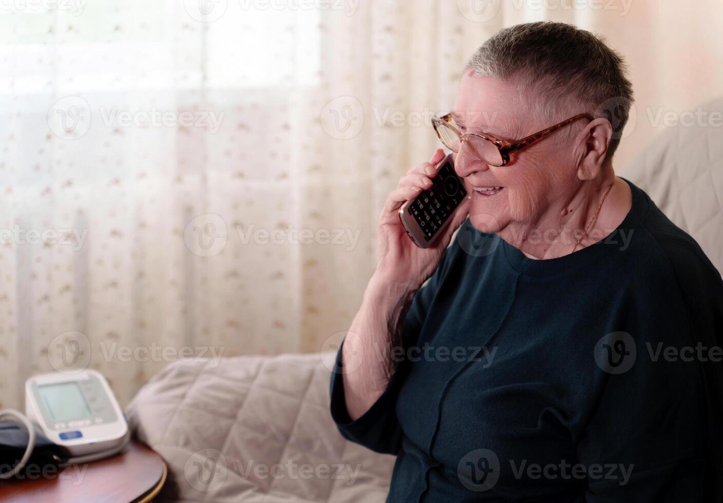 Retired senior engages in a phone conversation while a medical device for monitoring blood pressure sits nearby, demonstrating the integration of technology into daily life photo