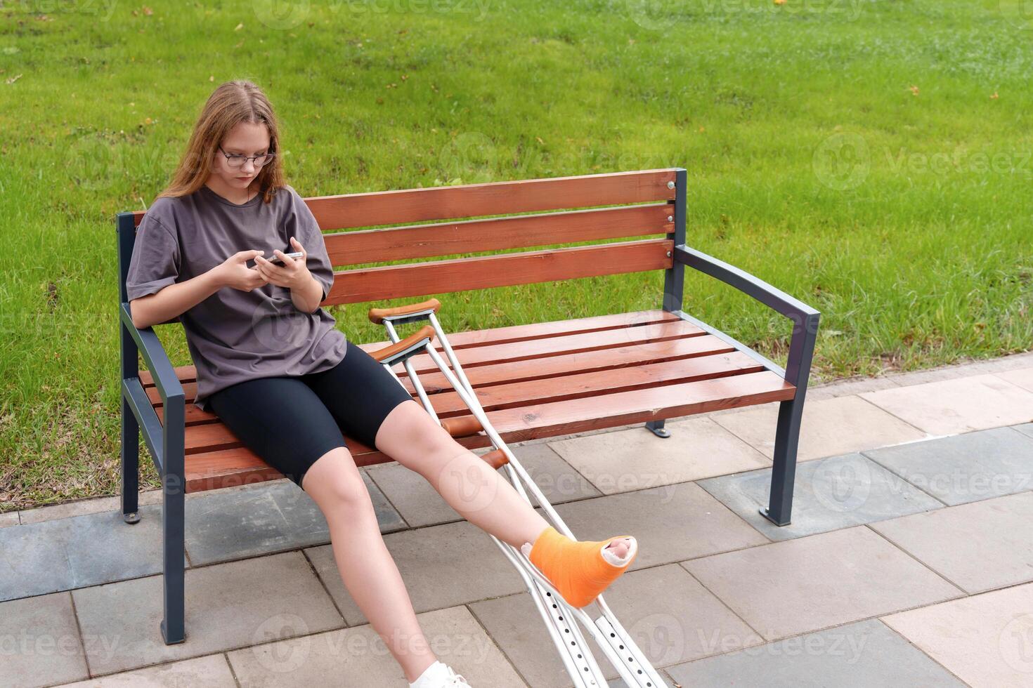 Teenage patient with a leg cast resting on a park bench, leveraging her smartphone for social interaction and calls during her rehabilitation journey in the peaceful summer setting photo