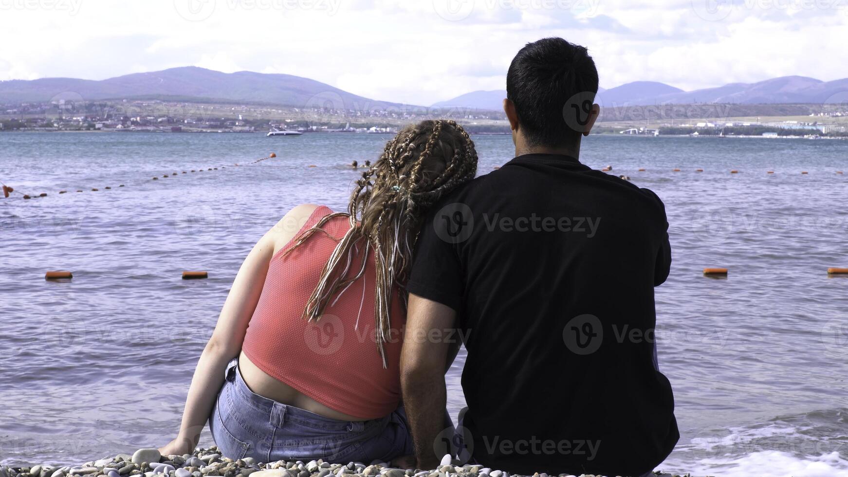 Rear view of loving couple sitting on stony shore in front of the sea. Media. Honeymoon concept, man and woman in love enjoying glass of red wine on the beach on a sunny summer day. photo