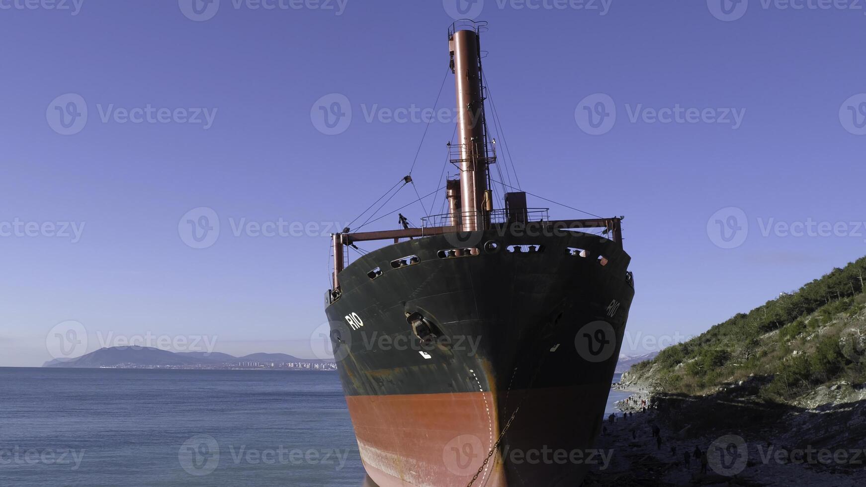 Aerial view of red industrial ship with people on board doing sailor work moored near sea shore. Shot. Big barge on the coastline near forested slope and people walking around. photo