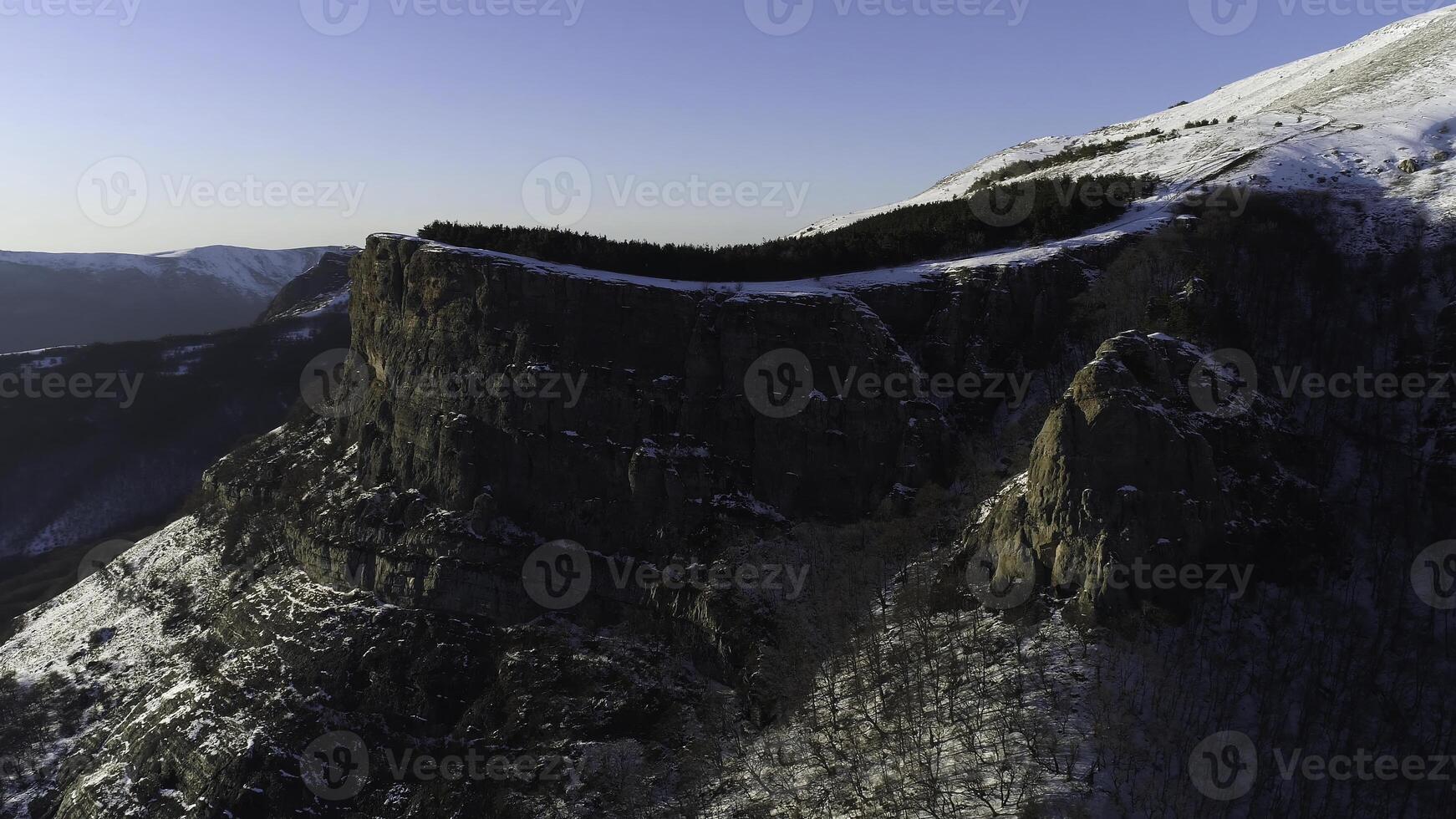 The slope of the high rock massif covered with pine trees on blue clear sky background. Shot. Aerial of the forested mountain slope and snowy peak. photo