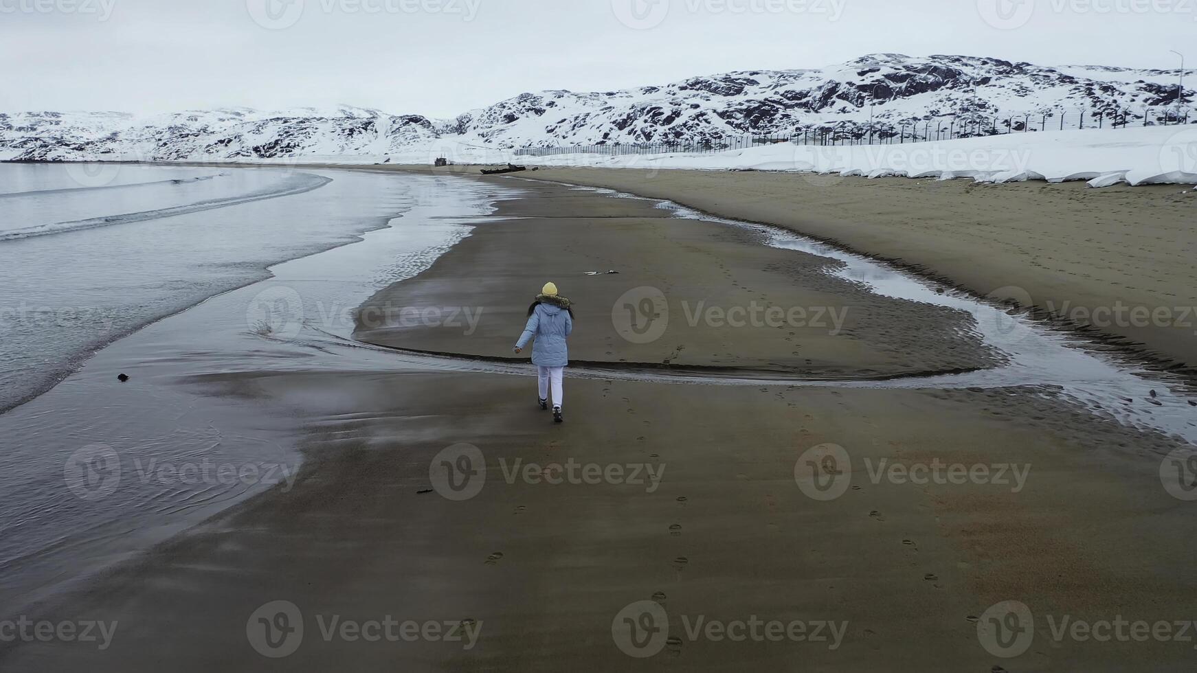 Aerial of a little girl in blue warm jacket runing along the coast of the cold winter sea. Footage. Rear view of a kid running on the sandy beach near snow covered hills. photo