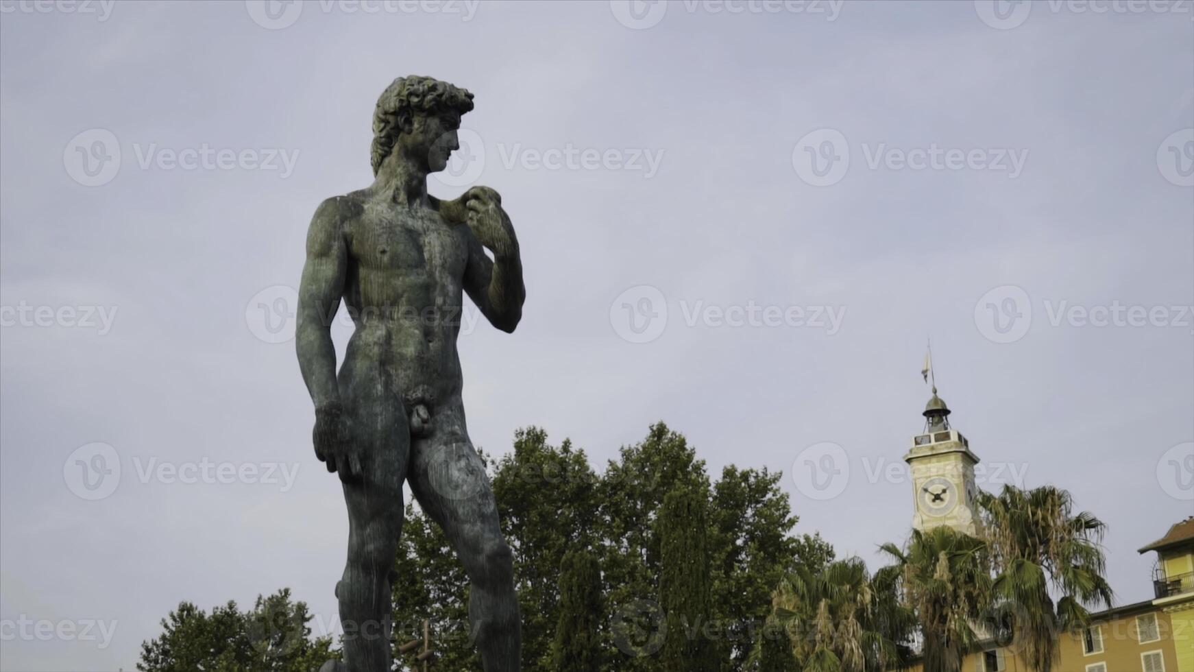 Statue of David in park. Action. Beautiful manly statue of David stands in park on background of sky. Nude statue of David is made of dark stone and stands on pedestal in park photo
