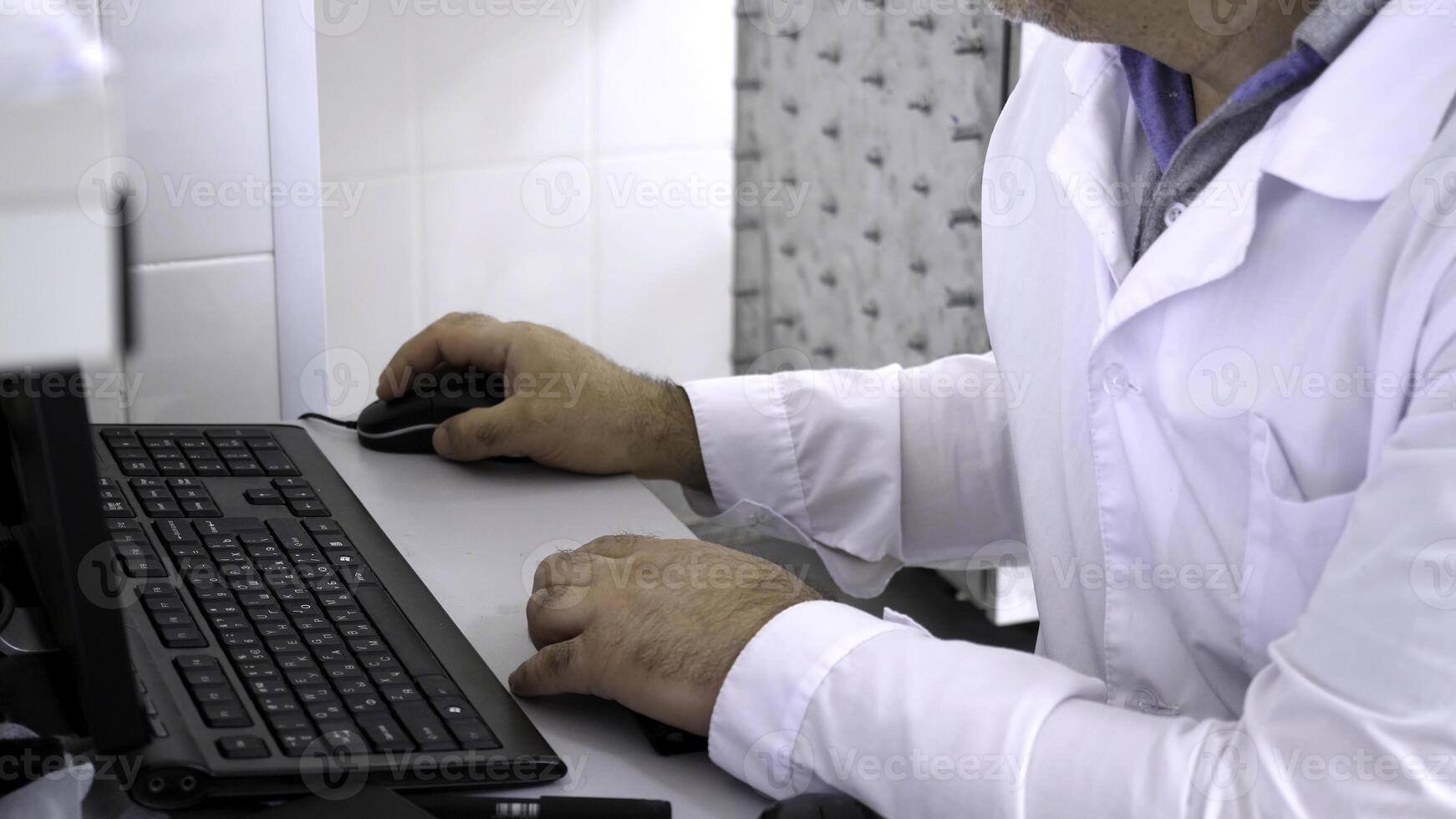 Close-up of man in white coat sitting at computer. Man in white coat sitting at computer in white room is doctor or scientist photo