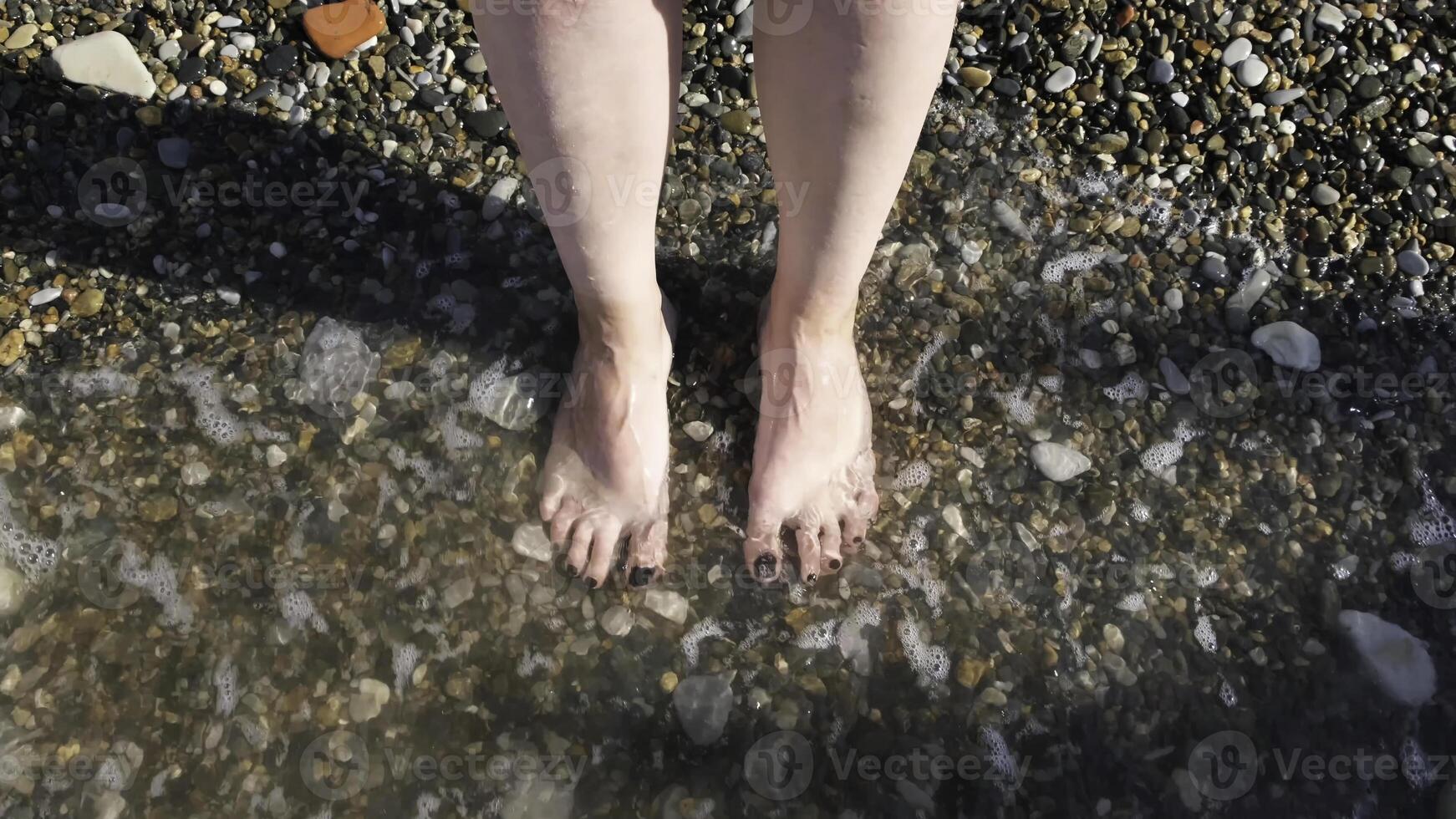 Top view of feet at the beach with sea waves. Concept. Connecting to nature, beautiful legs and feet of a woman with her nails painted black being washed by sea or ocean waves. photo