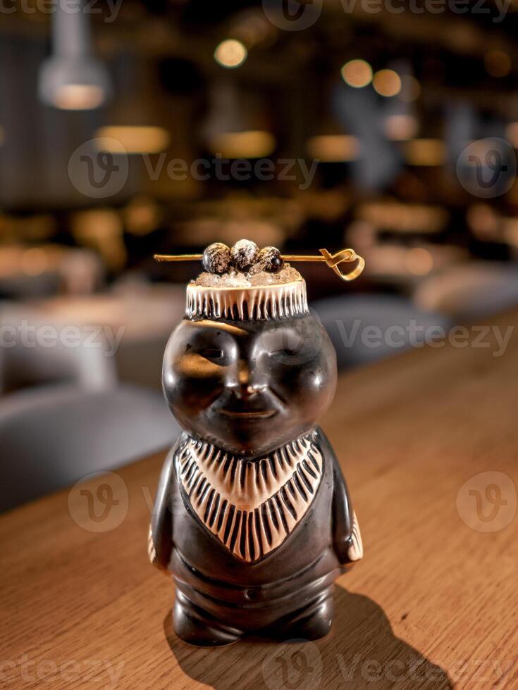 Authentic Tiki Drink Cocktail. Beautiful tropical tiki cocktail on bar counter. Tiki cocktail in monkey mug decorated fig and raspberry on bar in night club or restaurant interior photo