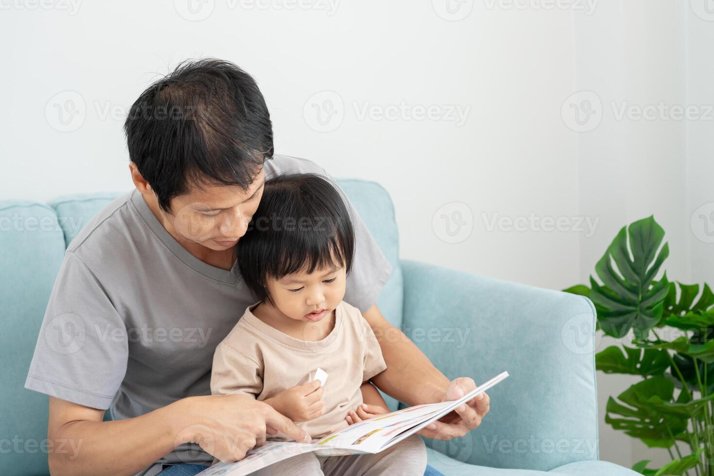Happy Asian father relax and read book with baby time together at home. parent sit on sofa with daughter and reading a story. learn development, childcare, laughing, education, storytelling, practice. photo