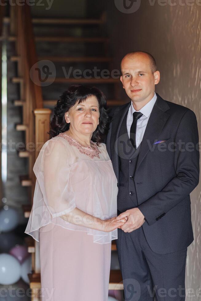 A man and a woman are standing in front of a wall. The man is wearing a suit and tie, and the woman is wearing a dress. They are both smiling and holding hands. Scene is happy and friendly photo