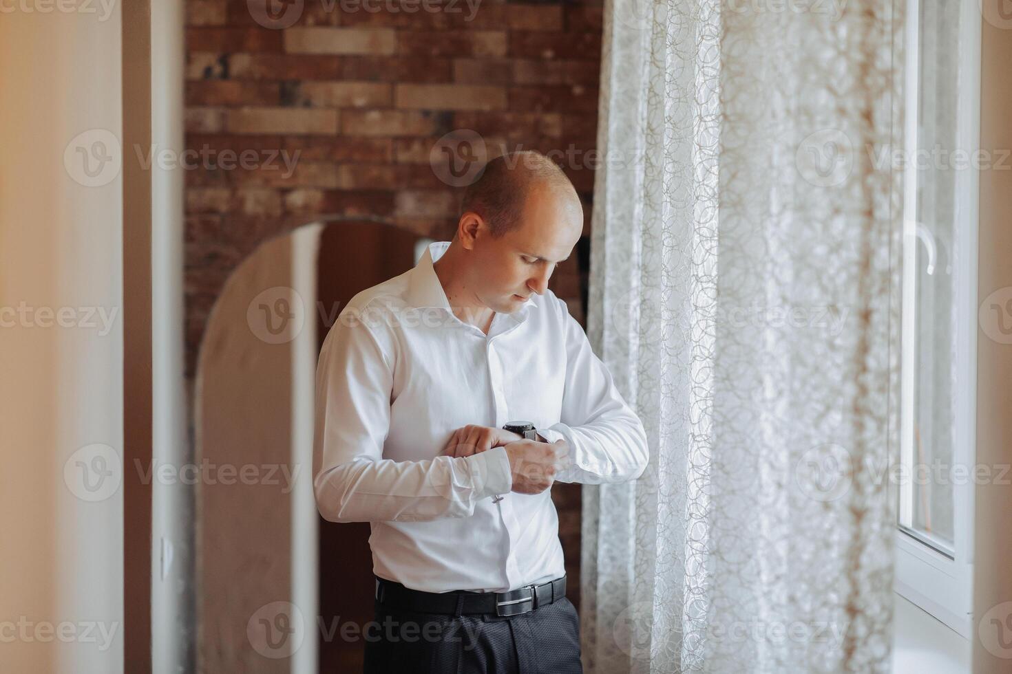 A man in a white shirt is getting dressed in front of a window. He is adjusting his shirt and looking out the window photo
