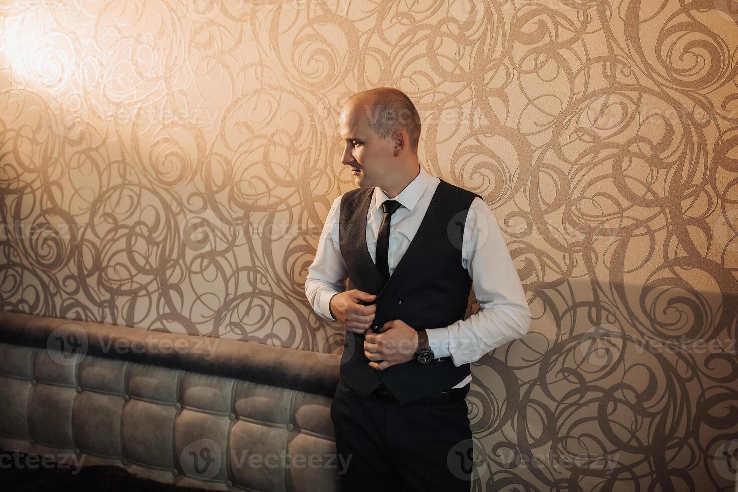 A man in a suit and vest stands in front of a wall with a patterned design. He is adjusting his tie and he is in a professional setting photo