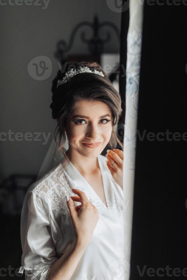 A woman in a white dress is smiling and looking out a window. The image has a warm and inviting mood, with the woman's smile and the light coming in through the window photo