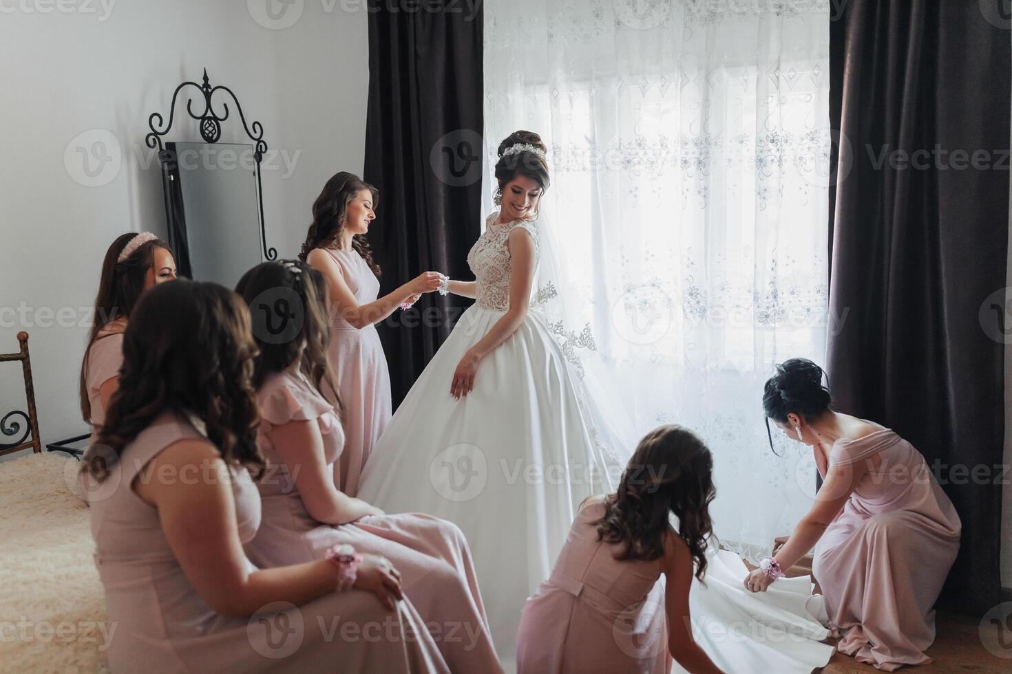 A bride and her bridesmaids are getting ready for the wedding. The bride is wearing a white dress and the bridesmaids are wearing pink dresses. They are all gathered in a room photo