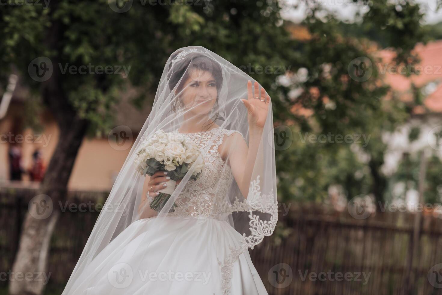 A woman in a white dress is holding a bouquet of flowers and is wearing a veil. She is smiling and she is happy photo