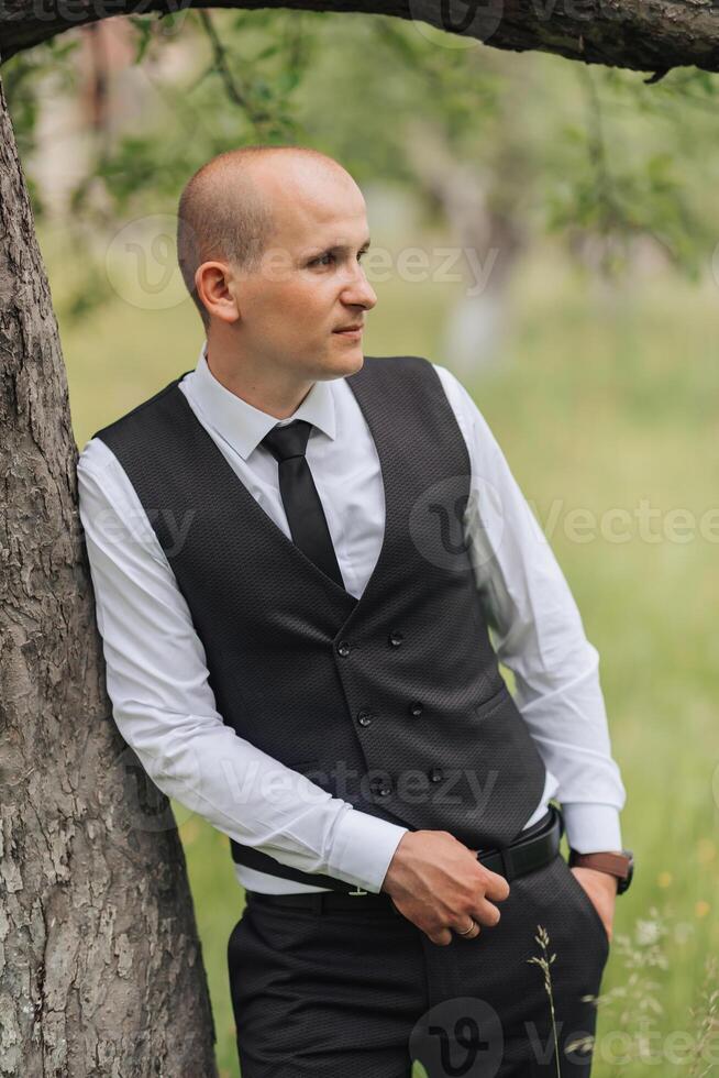 A man in a black vest and white shirt stands next to a tree. He is wearing a black tie and has his hands in his pockets. Concept of formality and elegance photo