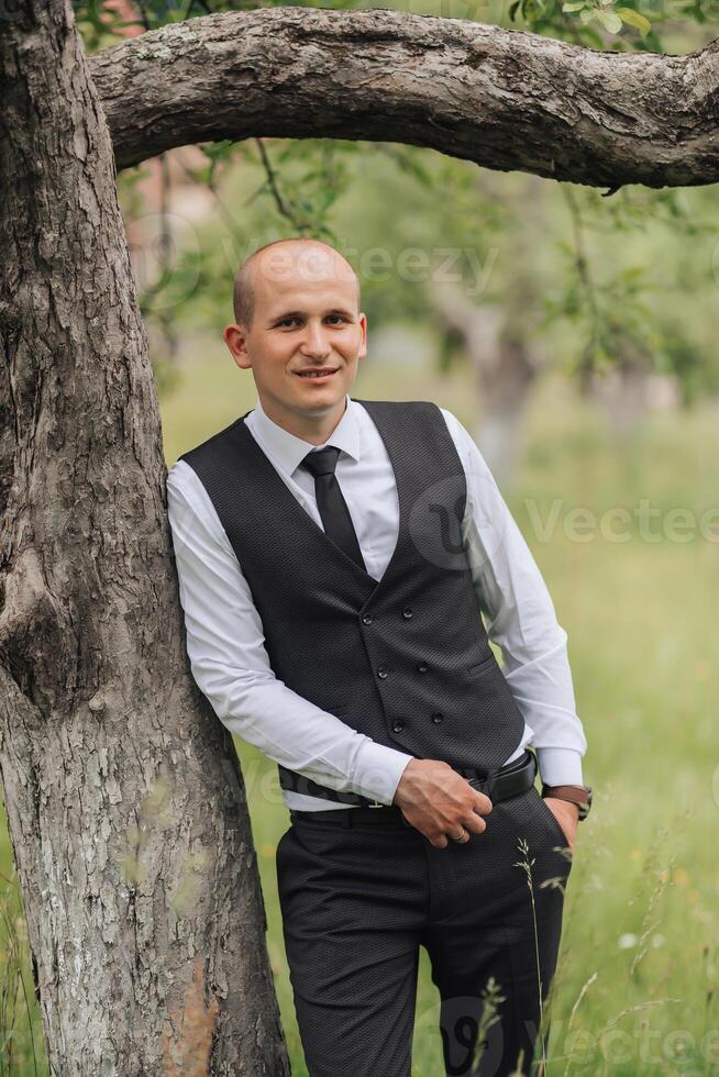 A man in a black vest and tie stands next to a tree. He is smiling and looking at the camera photo