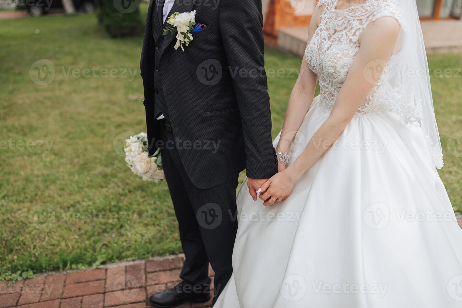 A bride and groom are standing together in a grassy area, holding hands. The bride is wearing a white dress and the groom is wearing a suit. The scene is likely a wedding or a special event photo