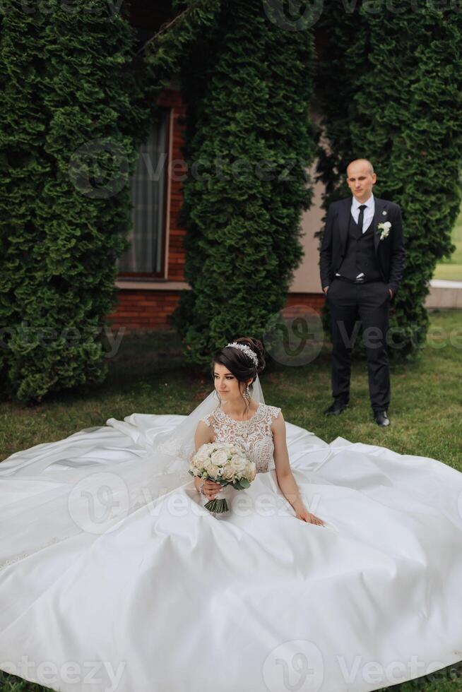 A bride and groom are standing in front of a large bush. The bride is wearing a white dress and holding a bouquet photo