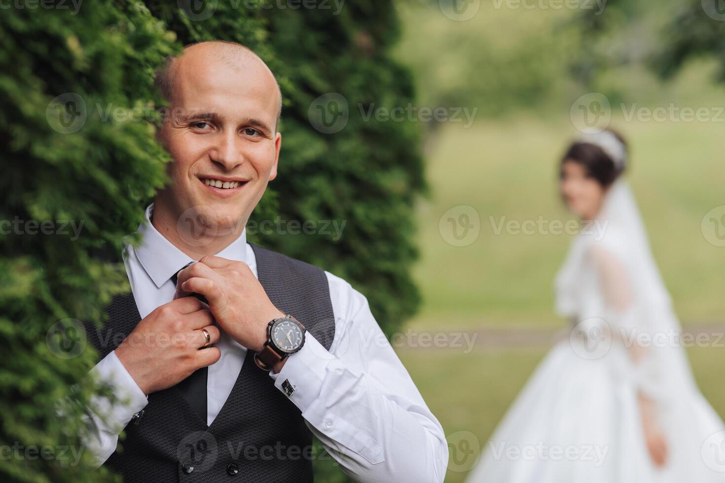 A man in a suit and vest is getting ready to tie his tie. A woman is standing behind him, wearing a white dress. Concept of formality and elegance, as the man is dressed for a special occasion photo