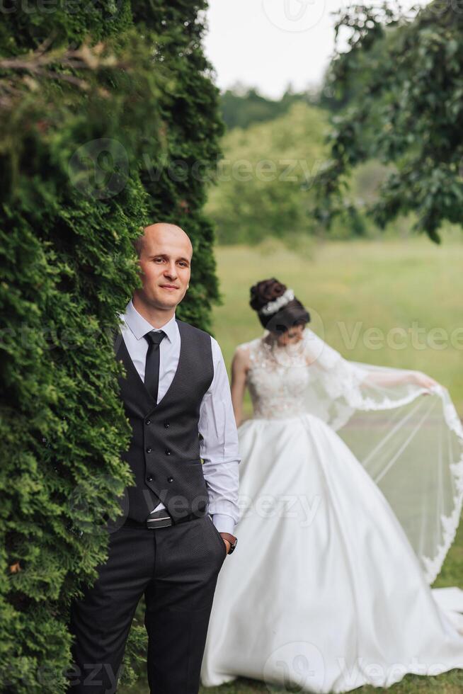 A man and a woman are posing for a picture in a field. The man is wearing a suit and tie, while the woman is wearing a white dress. They are standing in front of a bush photo