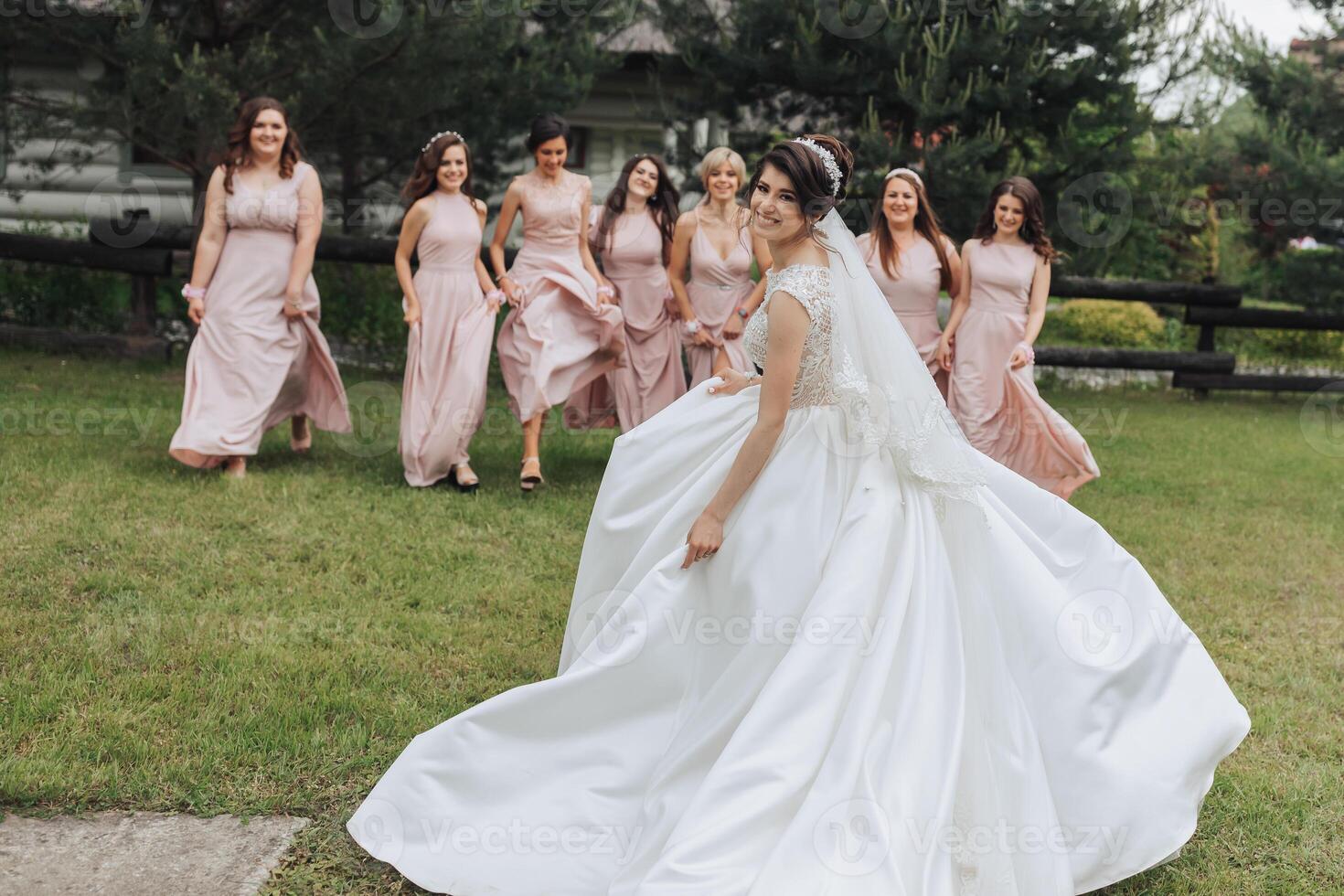 A bride in a white dress is surrounded by her bridesmaids. The bride is smiling and looking at the camera. The bridesmaids are all wearing pink dresses and are running around the bride photo