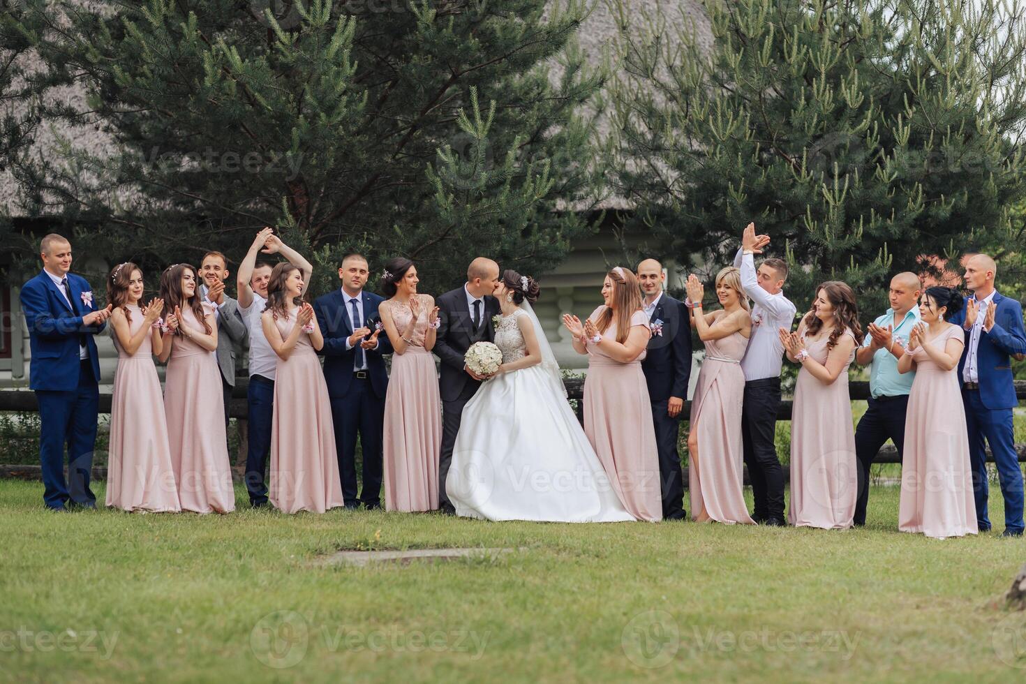 A group of people are gathered in a field, with a bride and groom in the center. The bride is wearing a white dress and the groom is wearing a suit photo